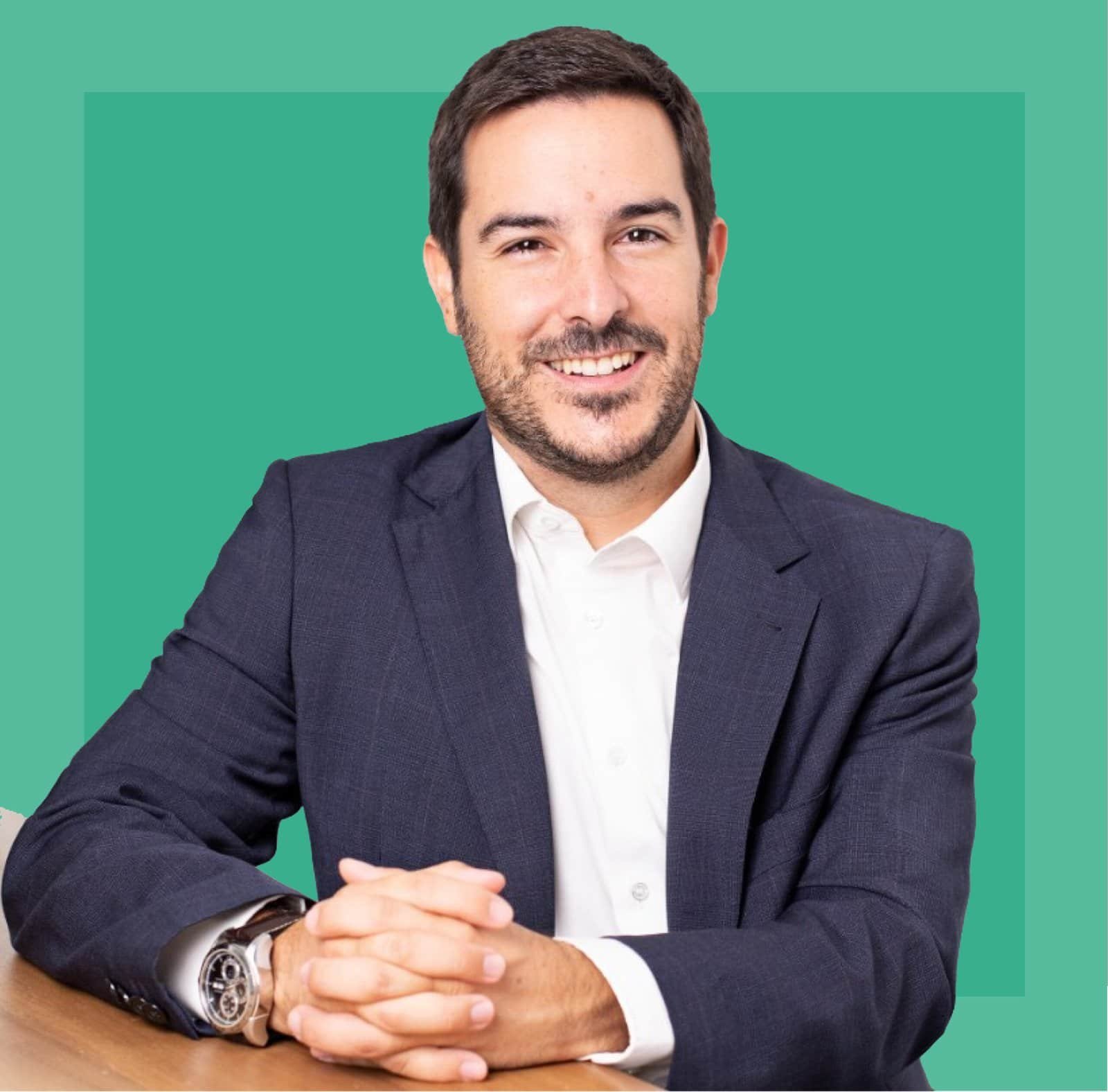 Manuel Cano sonriente con cabello corto y barba corta, vestido con un traje azul oscuro y camisa blanca, sentado con las manos entrelazadas sobre una mesa. Fondo de color verde sólido.